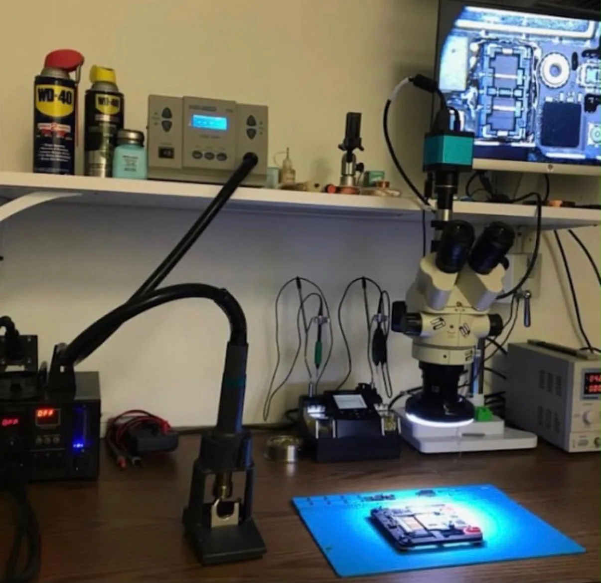 L'établi de réparation de la boutique : microscope binoculaire, station à air chaud, alimentation de laboratoire et carte mère de téléphone sous la loupe